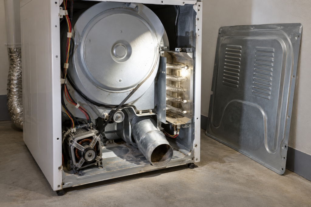 The rear of an electric dryer with its back panel removed and leaned against the wall, the coiled heating element visible inside its metal housing beside the exhaust duct, a flashlight beam from the side, no hands in frame.