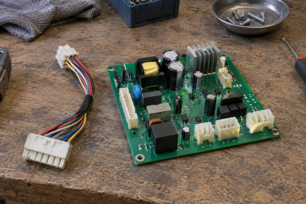 A washing machine motor control board lying on a workbench with its connector sockets visible and a loose wire harness beside it, soft overhead light.