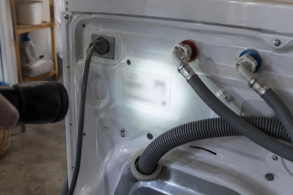 A white data plate on the rear panel of a washer with the water hoses and the power cord nearby, a flashlight beam from the side, the printed characters on the plate blurred and unreadable.